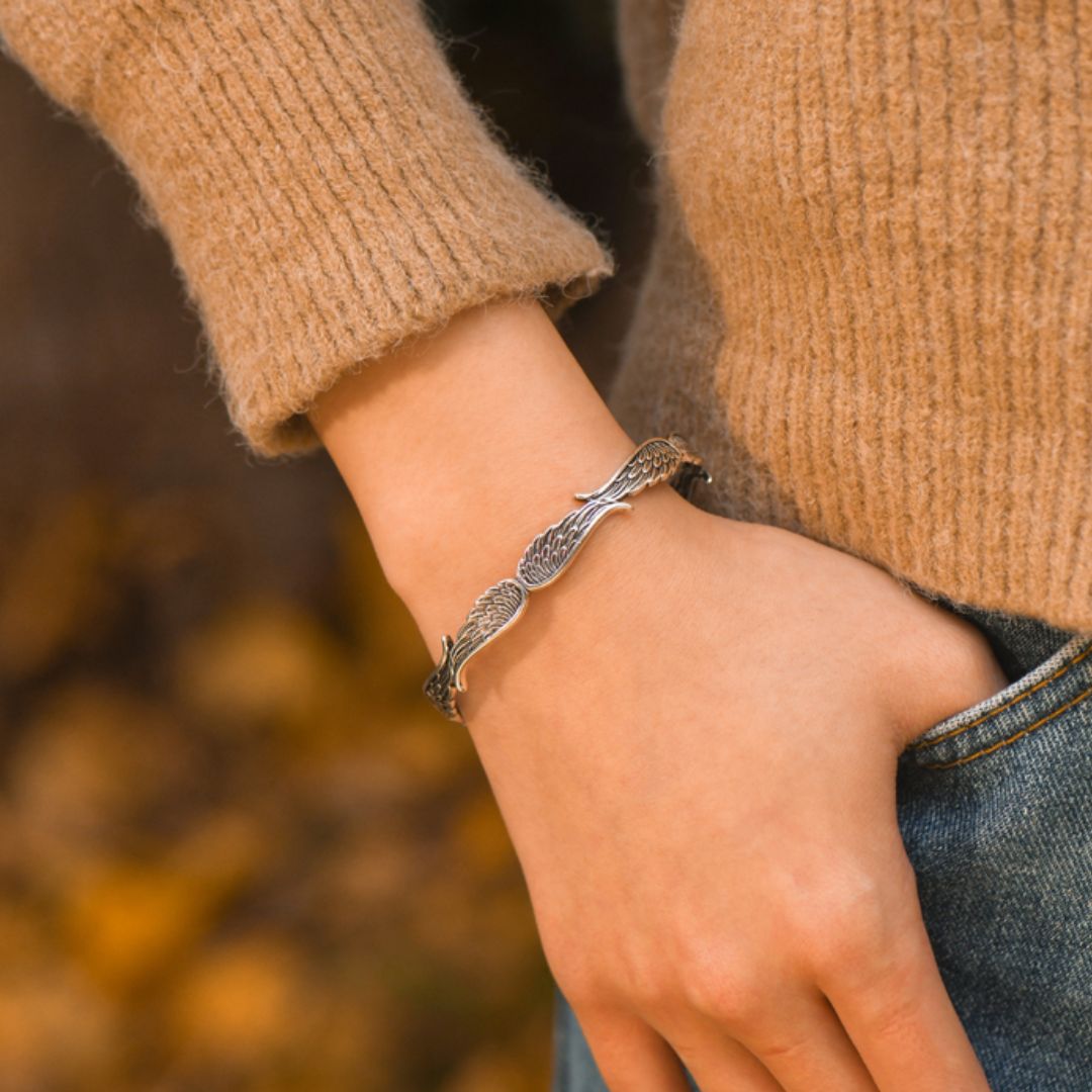 Lleva un jersey de punto marrón claro y vaqueros azules, la Pulsera Alas de ángel -una pulsera plateada en forma de pluma- en la muñeca y una mano en el bolsillo. El fondo borroso resplandece con cálidos colores otoñales.