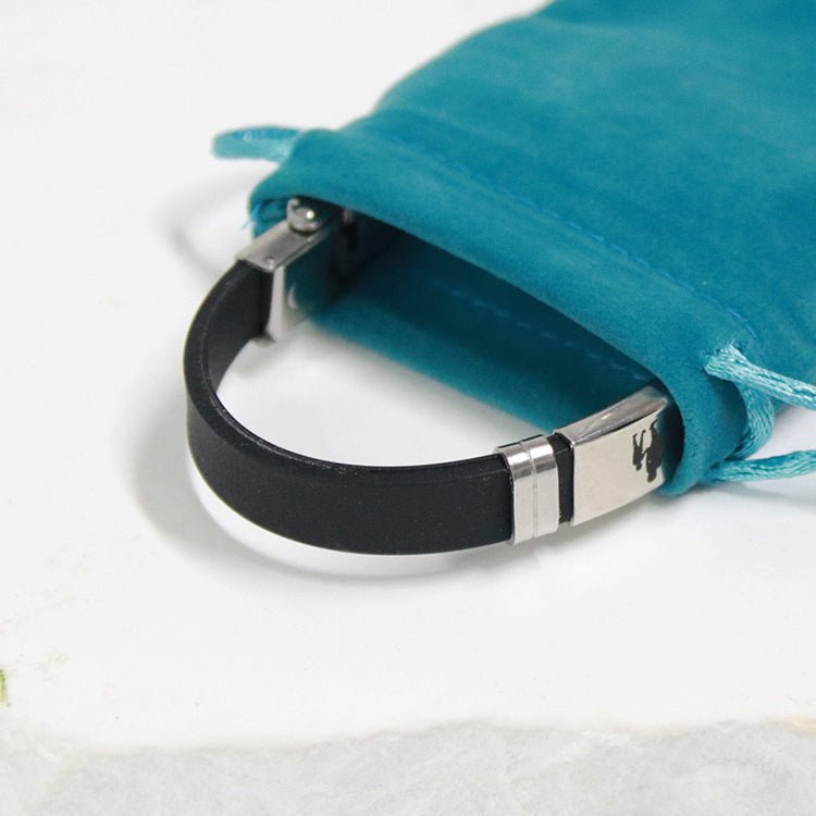 Un primer plano de la Pulsera con dedicatoria "Madre a hijo", una pulsera negra con cierre de acero inoxidable, parcialmente dentro de una bolsa de ante verde azulado sobre una superficie blanca.