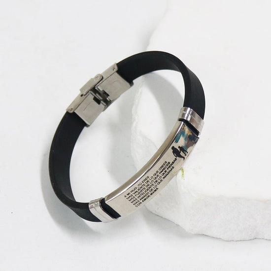 Pulsera de silicona negra con dedicatoria "Madre a hijo" con placa de acero inoxidable plateada, grabada con texto personalizado y un pequeño gráfico de una persona con un perro, sobre una superficie de piedra blanca. Ideal como regalo personalizado.