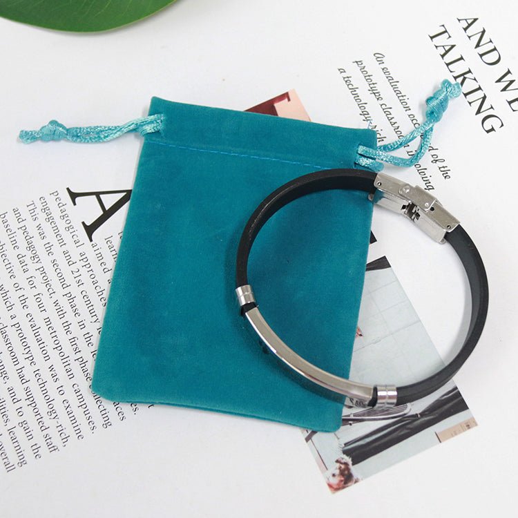 La pulsera con dedicatoria "Madre a hijo", en negro y plateado con cierre seguro, descansa sobre una bolsa azul de terciopelo sobre una revista. Fabricada en acero inoxidable, es un regalo personalizado y duradero para tu hijo.
