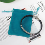 La pulsera con dedicatoria "Madre a hijo", en negro y plateado con cierre seguro, descansa sobre una bolsa azul de terciopelo sobre una revista. Fabricada en acero inoxidable, es un regalo personalizado y duradero para tu hijo.
