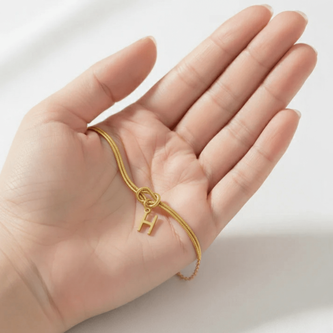 Una mano sostiene la Pulsera de nudo con inicial, con cadena dorada y colgante en forma de inicial, apoyada en la palma sobre un fondo suave y claro.