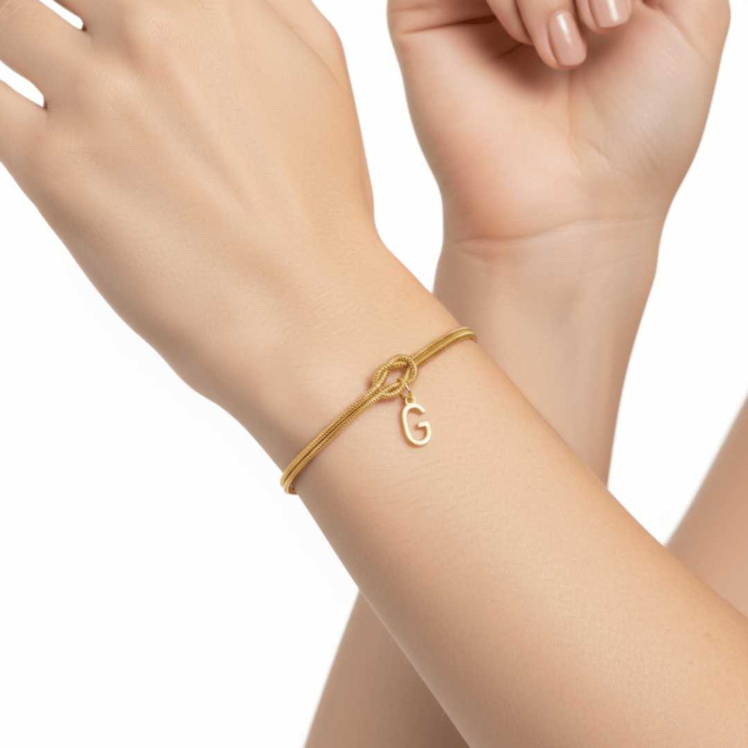 Primer plano de una muñeca que lleva la Pulsera de nudo con inicial, con un cordón dorado y una letra G dorada colgante. La otra mano toca suavemente el antebrazo, un regalo memorable que se muestra sobre un limpio fondo blanco.