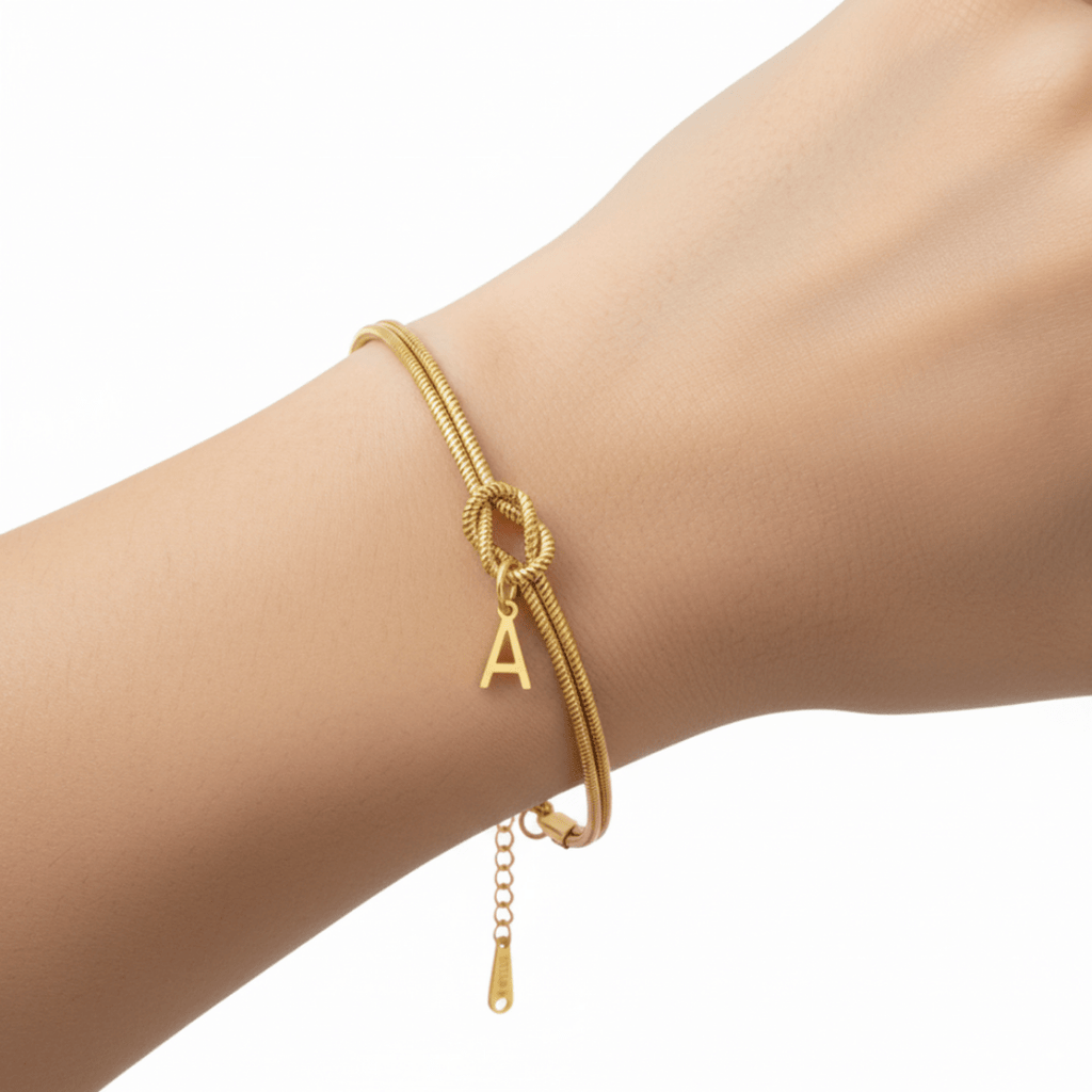 La Pulsera de nudo con inicial, que se muestra con un pequeño dije de la letra A en una muñeca sobre un fondo blanco liso, es un regalo inolvidable ideal.