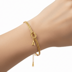 La Pulsera de nudo con inicial, que se muestra con un pequeño dije de la letra A en una muñeca sobre un fondo blanco liso, es un regalo inolvidable ideal.