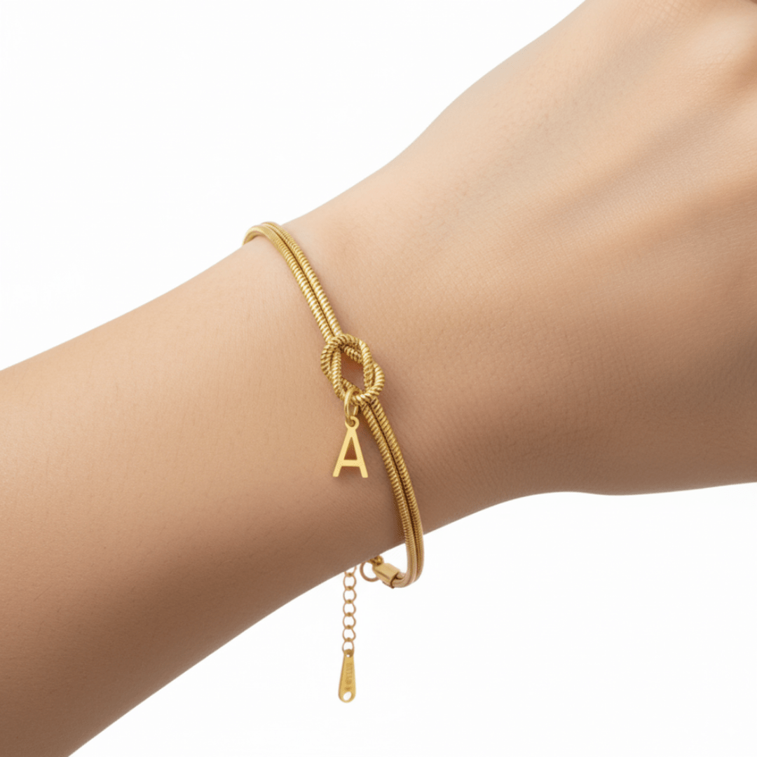 La Pulsera de nudo con inicial, que se muestra con un pequeño dije de la letra A en una muñeca sobre un fondo blanco liso, es un regalo inolvidable ideal.