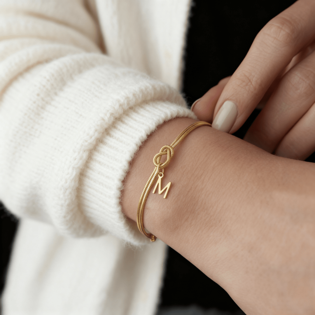 Una persona con jersey blanco se ajusta una Pulsera de nudo con inicial en la muñeca. La pulsera de oro, un regalo inolvidable, lleva un colgante con la letra M, y sus uñas claras están bien cuidadas.