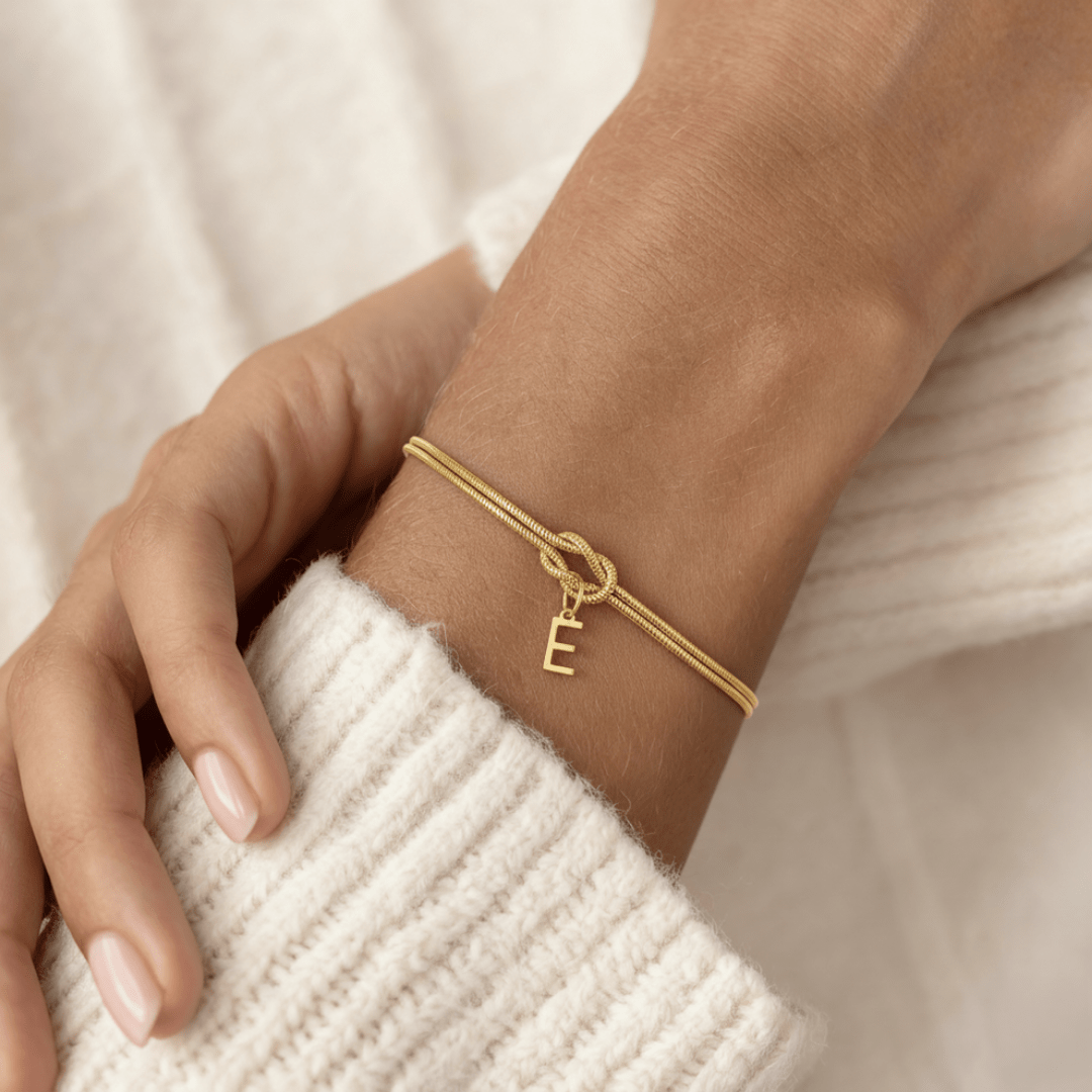 Un primer plano de una Pulsera de nudo con inicial -una elegante pulsera de oro con un dije E- descansa sobre la manga de un jersey de canalé color crema. Las uñas naturales y bien cuidadas hacen de esta pulsera personalizada un regalo memorable.