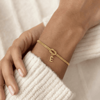 Un primer plano de una Pulsera de nudo con inicial -una elegante pulsera de oro con un dije E- descansa sobre la manga de un jersey de canalé color crema. Las uñas naturales y bien cuidadas hacen de esta pulsera personalizada un regalo memorable.