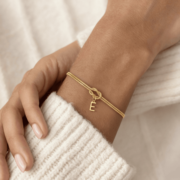 Un primer plano de una Pulsera de nudo con inicial -una elegante pulsera de oro con un dije E- descansa sobre la manga de un jersey de canalé color crema. Las uñas naturales y bien cuidadas hacen de esta pulsera personalizada un regalo memorable.