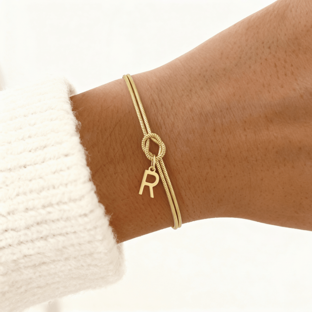 La muñeca de una persona luce una fina Pulsera de nudo con inicial, con un pequeño charm dorado en forma de letra R. Combinada con un jersey de punto blanco, una elegante pulsera personalizada y un regalo inolvidable.