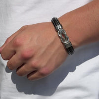 Una mano luce la pulsera con nudo infinito personalizable - A mi hijo, de cuero negro con detalles plateados y alambre, ideal como regalo para hijo, sobre una camisa blanca sencilla.