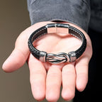 Una mano sostiene la "Pulsera con nudo infinito personalizable - A mi hijo", una pulsera de cuero trenzado negro con un nudo infinito de metal, perfecta como regalo para hijo o como pulsera de corazón madre e hijo.