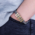 Una persona lleva una Pulsera de cadena personalizada -una pulsera de cadena de plata con dijes rectangulares de oro grabados con nombres- mientras apoya la mano en el bolsillo de unos vaqueros oscuros.