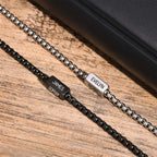 A pulsera de cubo personalizada en plateado, con los nombres "EVELYN" y "THOMAS" grabados, descansa junto a un artículo de cuero negro. Es el regalo perfecto para papá en el Día del Padre.
