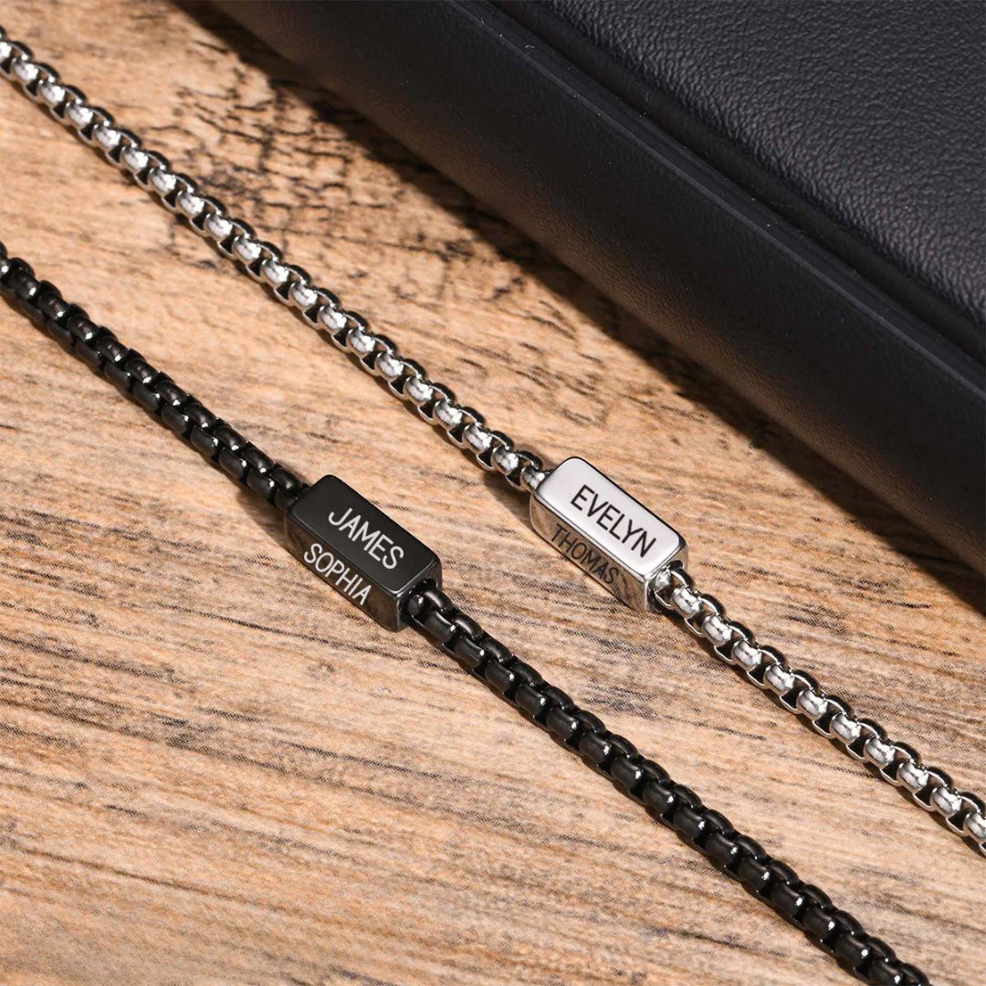 A pulsera de cubo personalizada en plateado, con los nombres "EVELYN" y "THOMAS" grabados, descansa junto a un artículo de cuero negro. Es el regalo perfecto para papá en el Día del Padre.