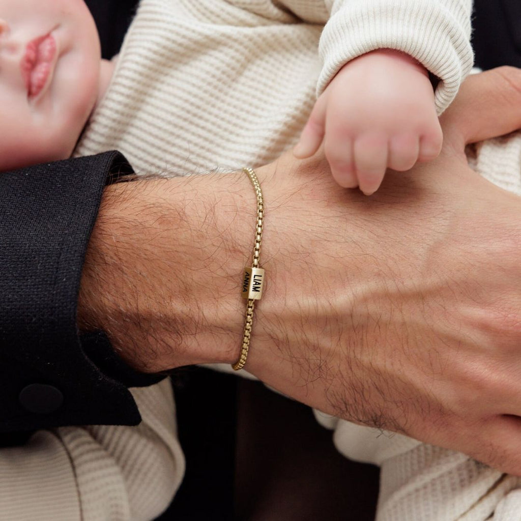 Primer plano de la mano de un adulto que lleva una Pulsera de cubo personalizada con un colgante cuadrado "MAMA", sujetando suavemente el brazo de un bebé vestido con un ligero conjunto de canalé: un momento conmovedor y un regalo perfecto Día del Padre.