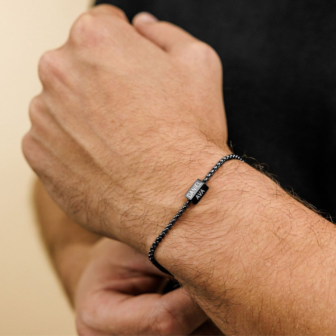 Detalle de una muñeca con la Pulsera de cubo personalizada en negro, de acero inoxidable, con dijes rectangulares grabados DANIEL y AMA. Ideal como regalo personalizado para papá o para el Día del Padre.