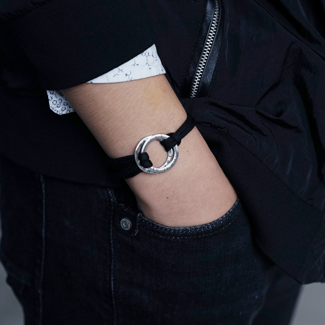 Una persona con jeans y chaqueta negros, camiseta blanca con pequeños estampados, lleva su mano en el bolsillo mostrando una Pulsera de cuerda para hombre grabada; un regalo ideal para él.