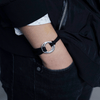 Una persona con jeans y chaqueta negros, camiseta blanca con pequeños estampados, lleva su mano en el bolsillo mostrando una Pulsera de cuerda para hombre grabada; un regalo ideal para él.
