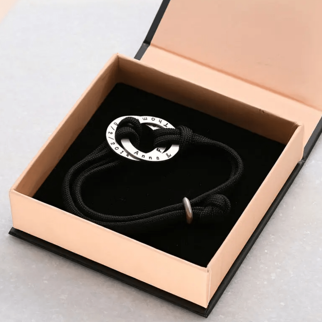 La Pulsera de cuerda para hombre grabada, con un colgante redondo de plata con nombres grabados, se presenta en un joyero de color melocotón con interior negro, perfecto como regalo para hombre.