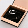La Pulsera de cuerda para hombre grabada, con un colgante redondo de plata con nombres grabados, se presenta en un joyero de color melocotón con interior negro, perfecto como regalo para hombre.