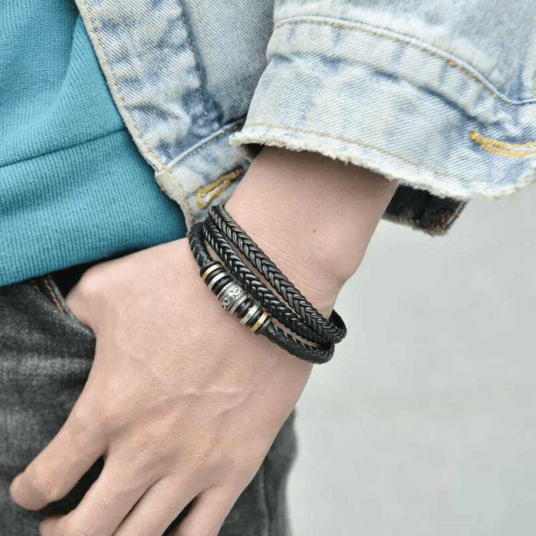 Primer plano de una muñeca que lleva la Pulsera de cuero - Al hombre de mi vida, de cuero trenzado negro con cuentas metálicas, combinada con una chaqueta vaquera azul y una camisa verde azulado, ideal como regalo amoroso o pulsera personalizada.