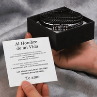 Una persona sostiene una nota en español junto a una caja de regalo negra abierta con la Pulsera de cuero - Al hombre de mi vida. Colocada sobre tela gris, es ideal como regalo amoroso o pulsera hombre.