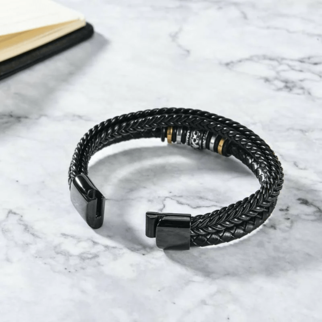 La Pulsera de cuero - Al hombre de mi vida, una pulsera de cuero trenzado negro con detalles metálicos, descansa sobre una superficie de mármol blanco cerca de la esquina de un cuaderno abierto.