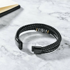 La Pulsera de cuero - Al hombre de mi vida, una pulsera de cuero trenzado negro con detalles metálicos, descansa sobre una superficie de mármol blanco cerca de la esquina de un cuaderno abierto.
