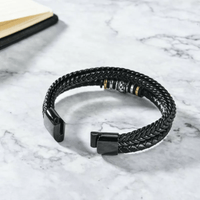 La Pulsera de cuero - Al hombre de mi vida, una pulsera de cuero trenzado negro con detalles metálicos, descansa sobre una superficie de mármol blanco cerca de la esquina de un cuaderno abierto.
