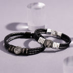 Dos pulseras de cuero negro trenzado con cierre plateado y charms grabados con nombres y fotos, ideales como pulsera de cuero con nombres y foto para hombre o un regalo original, exhibidas sobre una superficie gris clara.