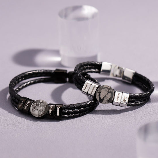Dos pulseras de cuero negro trenzado con cierre plateado y charms grabados con nombres y fotos, ideales como pulsera de cuero con nombres y foto para hombre o un regalo original, exhibidas sobre una superficie gris clara.