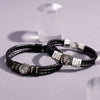 Dos pulseras de cuero negro trenzado con cierre plateado y charms grabados con nombres y fotos, ideales como pulsera de cuero con nombres y foto para hombre o un regalo original, exhibidas sobre una superficie gris clara.