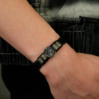 Un primer plano de la Pulsera de cuero con nombres y foto: una pulsera trenzada negra con cuentas de nombre grabadas y un charm redondo de foto. Combinada con una chaqueta oscura, es el regalo original perfecto para hombre.