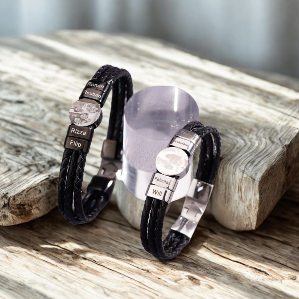 Dos pulseras de cuero trenzado negro "Pulsera de cuero con nombres y foto" con nombres e imágenes grabados en placas plateadas descansan sobre madera rústica, expuestas junto a un soporte cilíndrico transparente.