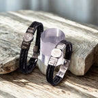 Dos pulseras de cuero trenzado negro "Pulsera de cuero con nombres y foto" con nombres e imágenes grabados en placas plateadas descansan sobre madera rústica, expuestas junto a un soporte cilíndrico transparente.