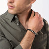 Un hombre con camisa verde abotonada se toca el cuello, lleva una Pulsera de cuero para Papá con trenzado negro y detalles plateados en la muñeca. Su cara es parcialmente visible, mirando hacia otro lado-ideal como regalo para hombre.