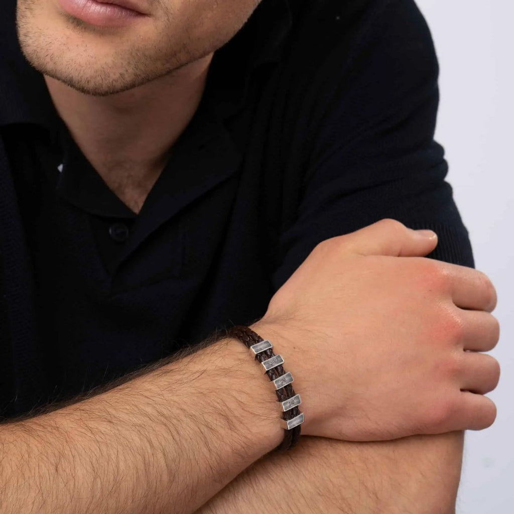 Un hombre vestido con un polo oscuro de manga corta apoya los brazos sobre una mesa y lleva la Pulsera de cuero para Papá con segmentos metálicos rectangulares en la muñeca izquierda, un regalo ideal para hombre. Se le ve parcialmente la cara.