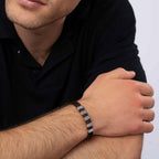 Un hombre vestido con un polo oscuro de manga corta apoya los brazos sobre una mesa y lleva la Pulsera de cuero para Papá con segmentos metálicos rectangulares en la muñeca izquierda, un regalo ideal para hombre. Se le ve parcialmente la cara.
