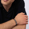 Un hombre vestido con un polo oscuro de manga corta apoya los brazos sobre una mesa y lleva la Pulsera de cuero para Papá con segmentos metálicos rectangulares en la muñeca izquierda, un regalo ideal para hombre. Se le ve parcialmente la cara.