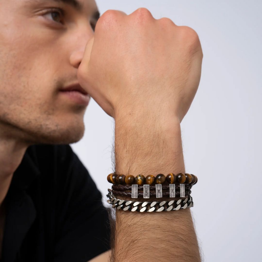 Un hombre lleva tres pulseras-una de cuentas marrones, otra de cuero trenzado con detalles metálicos, y la Pulsera de cuero para Papá personalizada con nombres-mientras reposa su barbilla en la mano. Ideal como regalo para hombre.