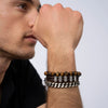 Un hombre lleva tres pulseras-una de cuentas marrones, otra de cuero trenzado con detalles metálicos, y la Pulsera de cuero para Papá personalizada con nombres-mientras reposa su barbilla en la mano. Ideal como regalo para hombre.
