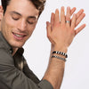 Un hombre sonriente de piel clara y pelo castaño corto lleva una camisa verde oscuro, un anillo de plata y una Pulsera de cuero para Papá, levantando las manos y mirando hacia abajo sobre un fondo claro-ideal como regalo para hombre.