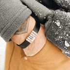 Una persona con una acogedora chaqueta de forro polar gris y pantalones marrones lleva una Pulsera de cuero personalizada con nombres, una pulsera de cuero trenzado negro con cuentas de plata grabadas con "Papá", "Mamá" y nombres, mientras los copos de nieve se posan suavemente sobre la chaqueta.