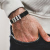 Primer plano de una muñeca que lleva la Pulsera de hombre con nombres grabados, con tres etiquetas metálicas rotuladas AMAZON, KOHLS y TIKTOK. La persona lleva un jersey blanco con textura y vaqueros oscuros: un regalo original ideal para hombre.