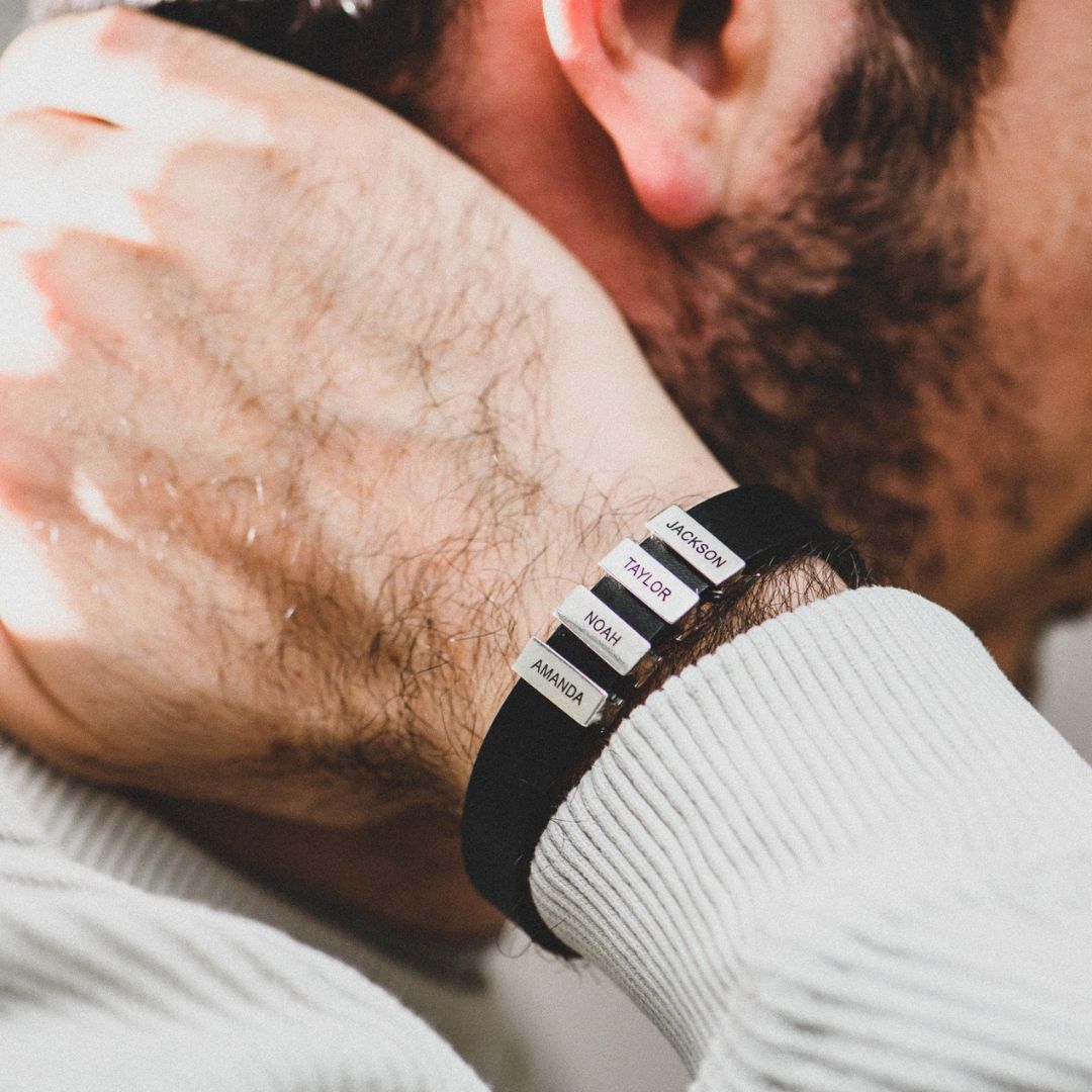 Primer plano de la muñeca de un hombre que lleva la Pulsera de hombre con nombres grabados en negro, con cuatro etiquetas grabadas con nombres -JACKSON, TAYLOR, MOM y AMANDA-, un regalo original ideal para el hombre que se toca la nuca.