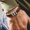 Un primer plano muestra una muñeca que lleva la Pulsera de hombre con nombres grabados, una pulsera de cuero marrón con tres placas de metal con nombres grabados con GRACE, JONATHAN y AUDREY, peinada con un jersey verde y unos vaqueros azules para un original look de regalo.
