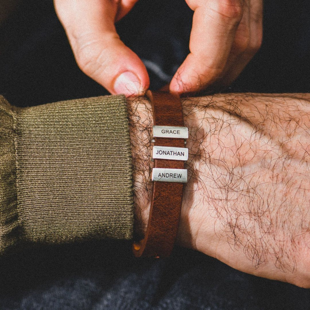 Una persona ajusta una Pulsera de hombre con nombres grabados -una pulsera de cuero marrón con tres etiquetas plateadas con nombres en las que se lee GRACE, JONATHAN y ANDREW- mostrando un regalo único y atento para hombres.