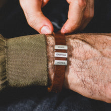 Cargar imagen en el visor de la galería, Una persona ajusta una Pulsera de hombre con nombres grabados -una pulsera de cuero marrón con tres etiquetas plateadas con nombres en las que se lee GRACE, JONATHAN y ANDREW- mostrando un regalo único y atento para hombres.